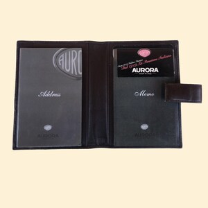 May include: A black leather address book with a memo pad and a clear plastic pocket for a business card. The business card has the text "Since 1919 - La Passione Italiana - AURORA - made in Italy".