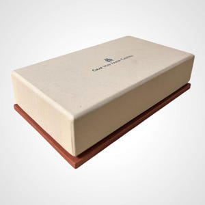 Puede incluir: Una caja rectangular con tapa beige claro y base marrón rojizo oscuro. La tapa tiene el texto "GRAF VON FABER-CASTELL" impreso en azul oscuro. La caja está sobre un fondo blanco.
