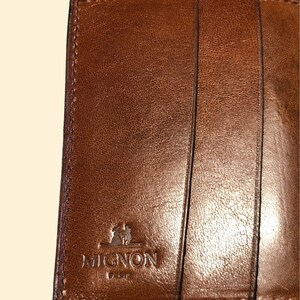May include: A brown leather wallet with a stitched edge and the embossed logo "MIGNON" in the lower center.