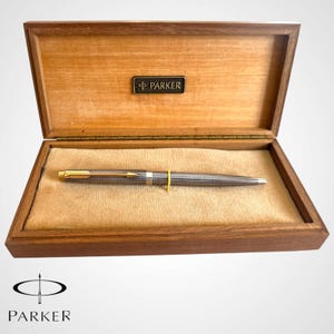 May include: A silver-toned ballpoint pen with a textured grid pattern and a gold-coloured arrow clip, resting inside a rectangular wooden presentation box. The interior of the lid features a black plaque with the text PARKER.