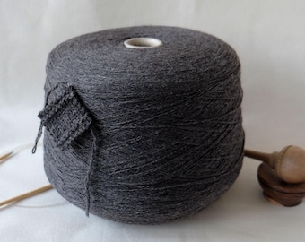 May include: A large spool of dark gray yarn with a small knitted swatch attached. Two wooden knitting needles and a wooden yarn swift are also present. The image has a soft, neutral background, and the text "Loopydot" is visible.