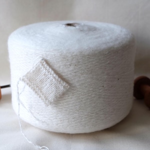 Puede incluir: Una madeja de hilo blanco con una pequeña muestra tejida adjunta. El hilo está enrollado alrededor de un tubo de cartón.