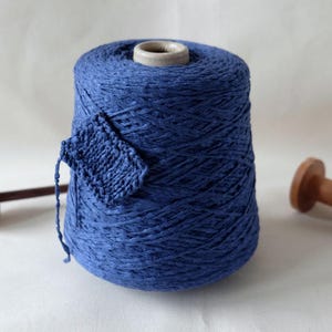 May include: A large spool of indigo blue yarn with a small knitted swatch attached. A wooden spindle and a wooden spool are on either side of the yarn. The yarn is for knitting and other fiber crafts.