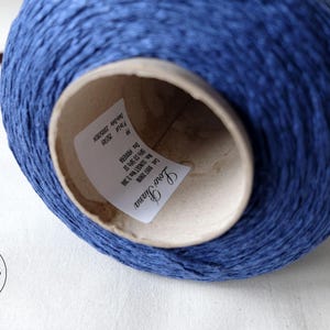May include: A close-up of a large spool of blue yarn. The yarn is tightly wound around a cardboard core. The label inside the core reads "Loro Piana". A wooden tool is visible in the background.