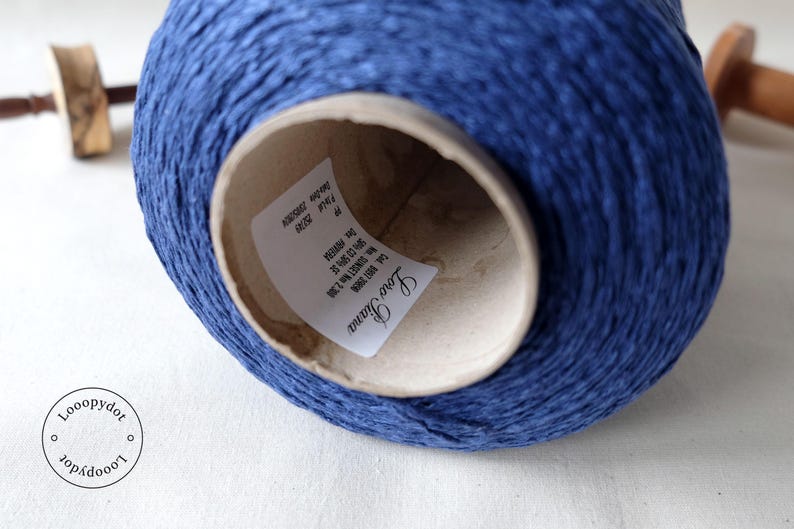May include: A close-up of a large spool of blue yarn. The yarn is tightly wound around a cardboard core. The label inside the core reads "Loro Piana". A wooden tool is visible in the background.