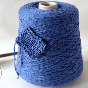 May include: A large spool of blue yarn with a small knitted swatch attached. A wooden spindle and a logo are also visible. The yarn is a vibrant shade of blue, and the knitted swatch shows the texture of the yarn.