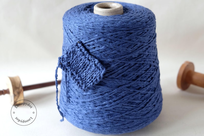 May include: A large spool of blue yarn with a small knitted swatch attached. A wooden spindle and a logo are also visible. The yarn is a vibrant shade of blue, and the knitted swatch shows the texture of the yarn.