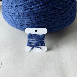May include: A close-up of a large ball of indigo blue yarn with a small spool of matching yarn in front. The small spool is white with indigo blue yarn wrapped around it. The background is a light-colored fabric.