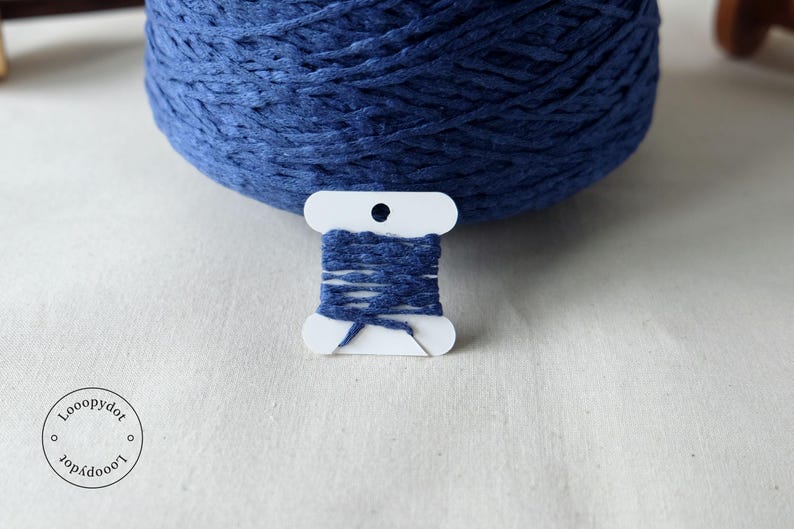 May include: A close-up of a large ball of indigo blue yarn with a small spool of matching yarn in front. The small spool is white with indigo blue yarn wrapped around it. The background is a light-colored fabric.