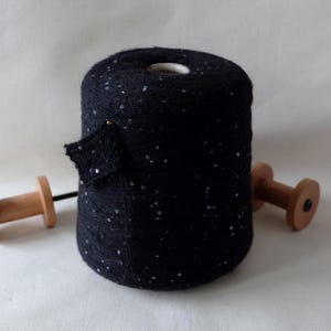 May include: A dark navy blue yarn cone with silver sequins. The yarn is cylindrical, with a small piece of the yarn pulled out. Two wooden spools are on either side of the yarn cone.