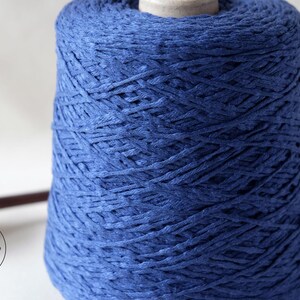 May include: A large spool of indigo blue yarn. The yarn is tightly wound, creating a textured surface. The spool sits on a white surface, with a wooden tool visible on the left side. The yarn appears to be made of a natural fiber.