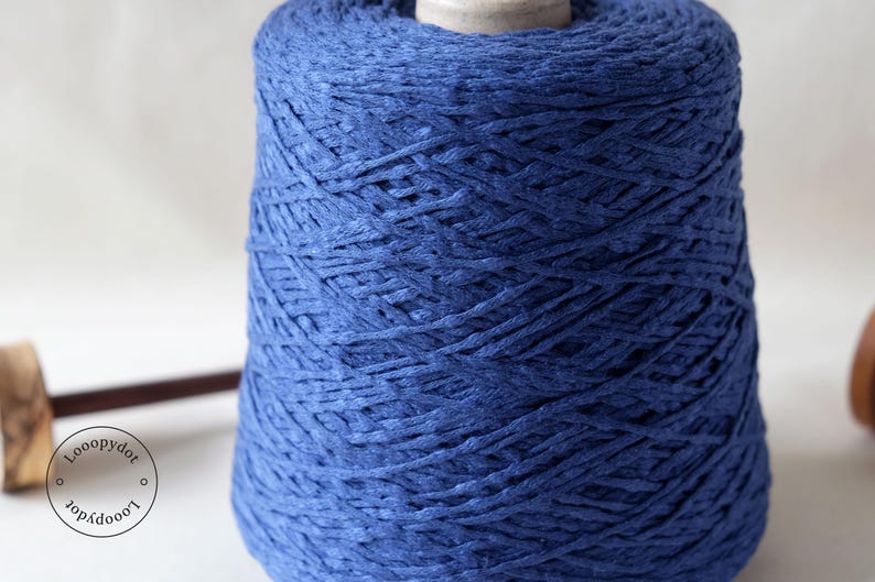 May include: A large spool of indigo blue yarn. The yarn is tightly wound, creating a textured surface. The spool sits on a white surface, with a wooden tool visible on the left side. The yarn appears to be made of a natural fiber.