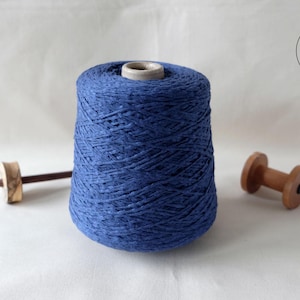 May include: A large cone of indigo blue yarn with a wooden spindle and a wooden spool. The yarn is tightly wound, and the wooden tools are light brown. The background is a plain white surface.