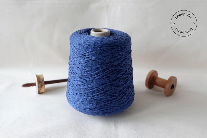 May include: A large cone of indigo blue yarn with a wooden spindle and a wooden spool. The yarn is tightly wound, and the wooden tools are light brown. The background is a plain white surface.