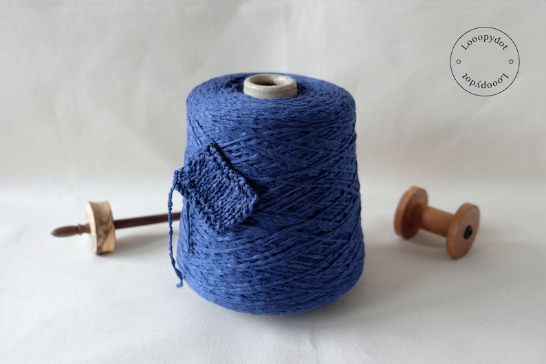May include: A large spool of indigo blue yarn with a small knitted swatch attached. A wooden spindle and a wooden spool are on either side of the yarn. The yarn is for knitting and other fiber crafts.