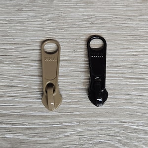 May include: Two metal zipper pulls, one tan and one black, are displayed against a light wood grain background. Each pull has a ring at the top and a zipper slider at the bottom. The brand name "YKK" is visible on each pull.