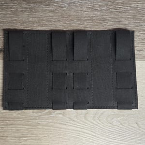 May include: Black rectangular MOLLE panel with multiple vertical and horizontal straps. The panel is made of a durable, textured fabric and is designed for attaching pouches and accessories. The panel is shown on a light wood surface.