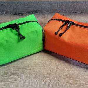 May include: Two rectangular zippered pouches, one bright green and one orange, sit side-by-side. Each has a black zipper and a black handle. The pouches are made of a textured fabric and rest on a wooden surface.