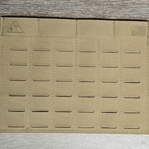 May include: Tan MOLLE panel with multiple horizontal slots for attaching gear. The panel has a rectangular shape and is made of a durable fabric. The top left corner features a mountain logo. The panel is designed for tactical or outdoor use.