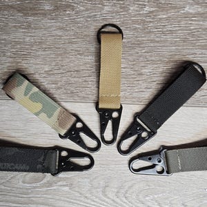 May include: Five keychains with different colored straps and black metal clips. The straps are camouflage, solid black, solid tan, solid green, and solid dark gray.