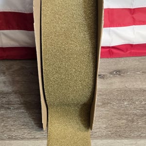 May include: A roll of olive green hook and loop fastener tape, also known as Velcro, is displayed in a cardboard dispenser. The American flag is visible in the background, with the tape unrolling onto a wooden surface.