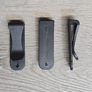 May include: Three black plastic clips of varying designs are arranged on a light wood-grain surface. The clips include a curved clip, a rectangular clip, and a simple spring clip. Each clip has a small hole.