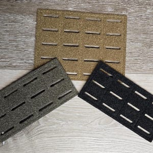 May include: Three rectangular panels featuring a grid of horizontal laser-cut slots. One panel is tan, one is olive green, and one is black. Each panel has a textured surface and is arranged in a fan-like formation on a flat surface.