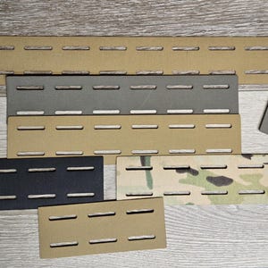 May include: A collection of various-sized tan, gray, black, and camouflage-patterned panels with horizontal slotted cutouts. The panels are arranged on a light wood-grain surface, showcasing their different shapes and colors.