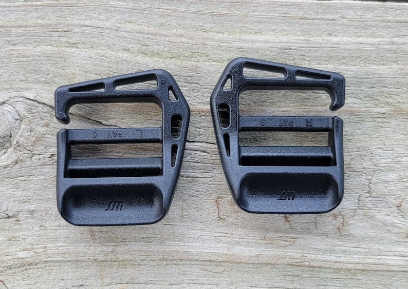 May include: Two black plastic buckles with a 'L PAT' and 'R PAT' marking on each. The buckles are designed for use with straps or webbing.