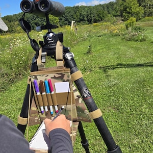 May include: A black tripod with attached binoculars, a camouflage pouch with pens, and a notepad. The tripod is set up outdoors on a grassy field under a blue sky. The binoculars are pointed towards the horizon.