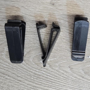 May include: Three black plastic belt clips are arranged on a light wood-grain surface. The clips are of different designs, including a spring-loaded clip and two other styles. The clips are designed to attach to a belt or other material.
