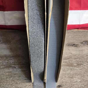 May include: Two rolls of gray hook and loop tape, one with a fuzzy surface and the other with a textured surface, are displayed. The rolls are in cardboard packaging, with the tape extending downwards. The background features an American flag.