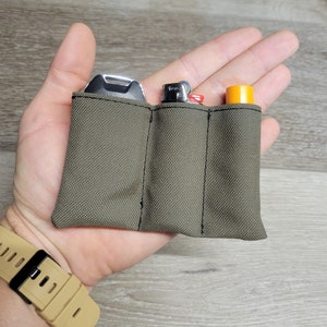 May include: A small, olive green pouch with three separate compartments. The pouch is made of a durable fabric and has black stitching. It is holding a silver multi-tool, a silver lighter, and a yellow tube.