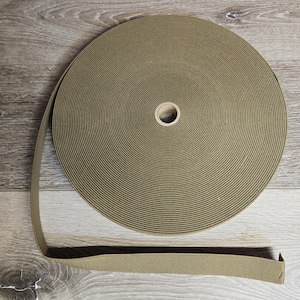 May include: A roll of olive green elastic band. The elastic is spooled around a cardboard core. A length of the elastic band extends from the roll, showing its texture and color. The background is a light wood grain.