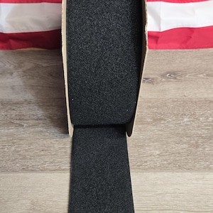May include: A roll of black, textured material, possibly felt or fabric, is partially unrolled, revealing a rectangular strip. The background features an American flag, with red and white stripes. The material is on a wooden surface.