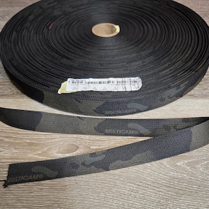 May include: A roll of black and green camouflage webbing. A length of the same webbing is unrolled and lies flat. The webbing has a textured surface and is suitable for various crafting and tactical applications.
