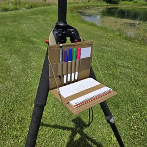 May include: A tan-colored field sketching kit attached to a tripod. The kit includes a notepad, several colored pens, and a pencil. The tripod is set up outdoors in a grassy area with a pond in the background.