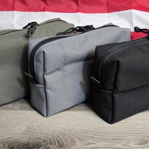 May include: Three rectangular zippered pouches in shades of green, gray, and black. Each pouch has a black zipper and a small loop on one side. The background features a wooden surface and an American flag.