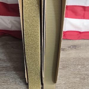 May include: Two rolls of hook and loop fastener tape, one with a fuzzy surface and the other with a looped surface. The tape is a tan color and is displayed in cardboard packaging. The background features an American flag.
