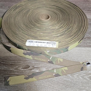 May include: A roll of camouflage webbing with a "MULTICAM" pattern. The webbing is tan, green, and brown. A section of the webbing is unrolled, showing a metal buckle.