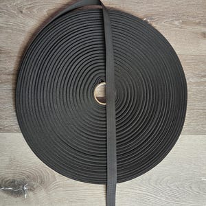 May include: A large roll of black elastic ribbon. The ribbon is tightly wound in a circular pattern, with a single length of ribbon extending from the roll. The ribbon is made of a woven material.