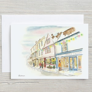 May include: Watercolor painting of a street scene with buildings and shops. The storefront of Bridge Cycles is visible. The artwork is on a white card with a white envelope. The artist's signature is in the lower left corner.