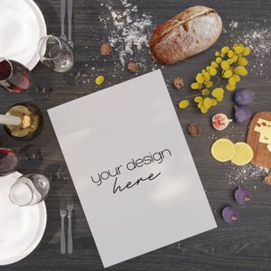 May include: A white blank card with the text "your design here" on a dark wood table with a loaf of bread, grapes, figs, lemons, and a candle.