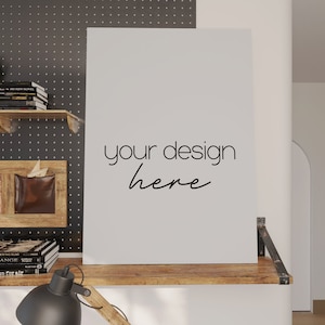 Puede incluir: Una maqueta de lienzo blanco con el texto "your design here" en un estante de madera con una lámpara negra y libros. El estante está fijado a una pared blanca con un tablero de clavijas negro detrás.