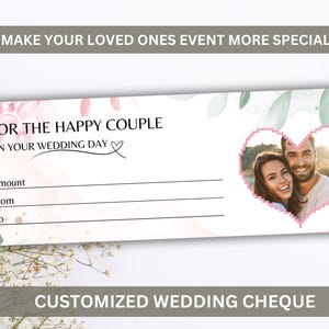 Puede incluir: Un cheque de boda personalizado con la foto de una pareja en un marco en forma de corazón rosa. El cheque tiene detalles florales y de hojas, con el texto "Para la feliz pareja en el día de su boda."