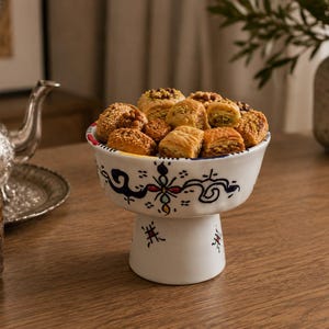 May include: A white ceramic pedestal bowl featuring hand-painted blue, red, and black decorative patterns, filled with an assortment of golden-brown, flaky baklava pastries topped with crushed nuts and walnuts, presented on a wooden surface.