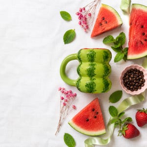 Pode incluir: Uma caneca de cerâmica texturizada verde-clara em forma de melancia, rodeada por fatias de melancia fresca, grãos de café escuros numa pequena taça rosa, ramos de flores cor-de-rosa, folhas de hortelã verde e dois morangos vermelhos.