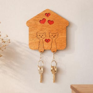 May include: Wooden key holder shaped like a house, featuring two cat silhouettes and red heart cutouts. Two sets of keys hang from the cat figures. The key holder is mounted on a light-colored wall, with a vase of dried flowers visible.