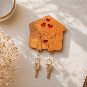 May include: Wooden key holder shaped like a house, featuring two cat silhouettes and three heart cutouts. Two keys hang from the bottom. The key holder is made of light brown wood and sits on a white surface.