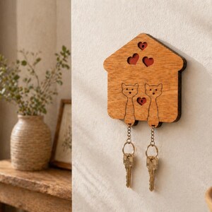 May include: Wooden key holder shaped like a house, featuring two cat silhouettes and heart cutouts. The key holder is made of light brown wood and has two keychains with keys attached. A decorative item for home organization.